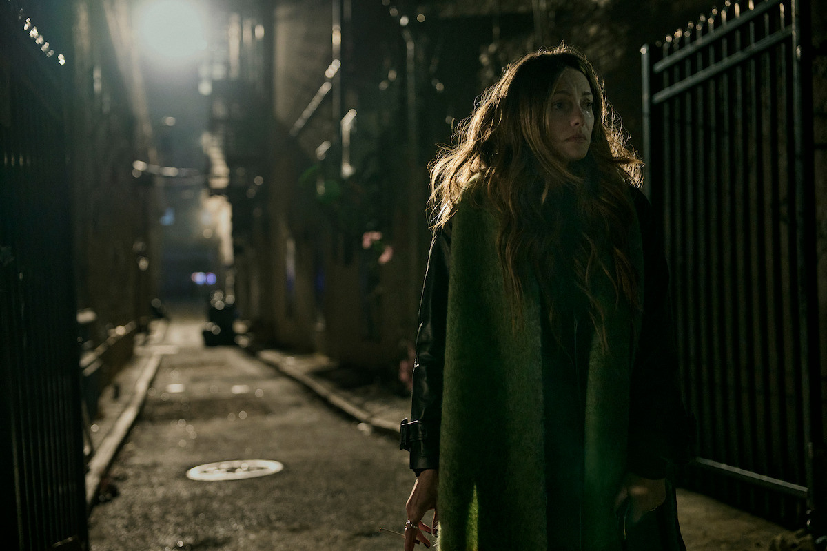 A woman with long hair stands in a dimly lit alleyway at night, surrounded by iron fences and shadows, creating a tense, mysterious atmosphere.