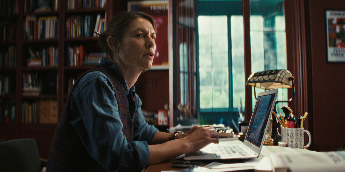 A woman sits at a desk in a cozy home office filled with books, working on a laptop. The room has warm lighting and large windows, creating a focused and intellectual mood.