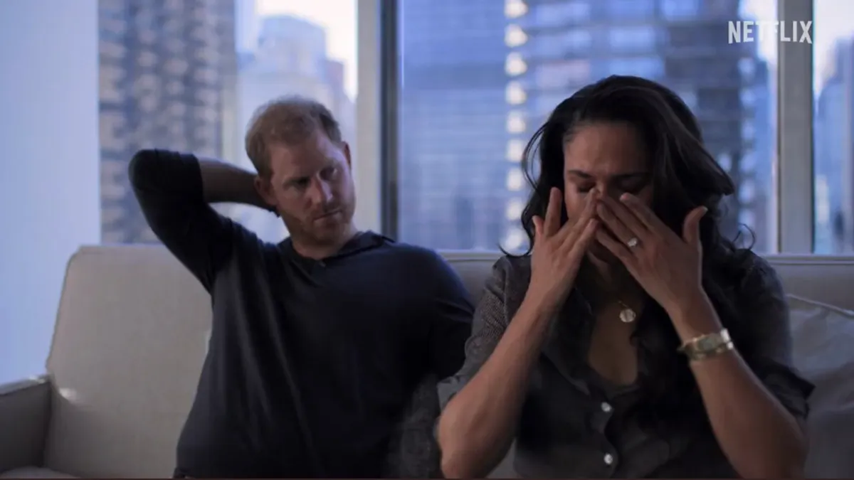 Meghan Markle cries on a couch and puts her hands to her face as Prince Harry with his arm behind his neck looks at her