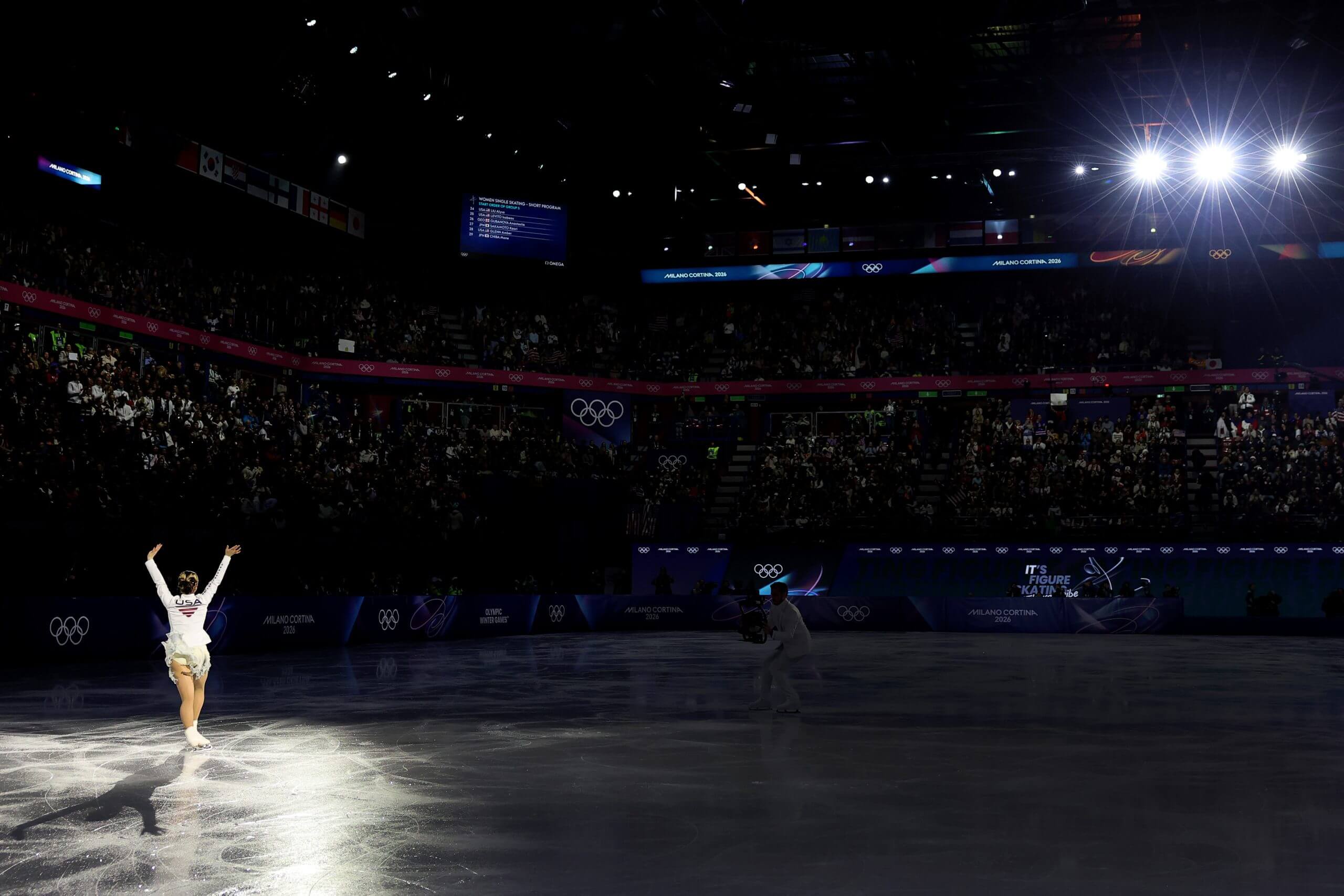 A shot of Alysa Liu from behind as she waves to the crowd in a darkened arena.