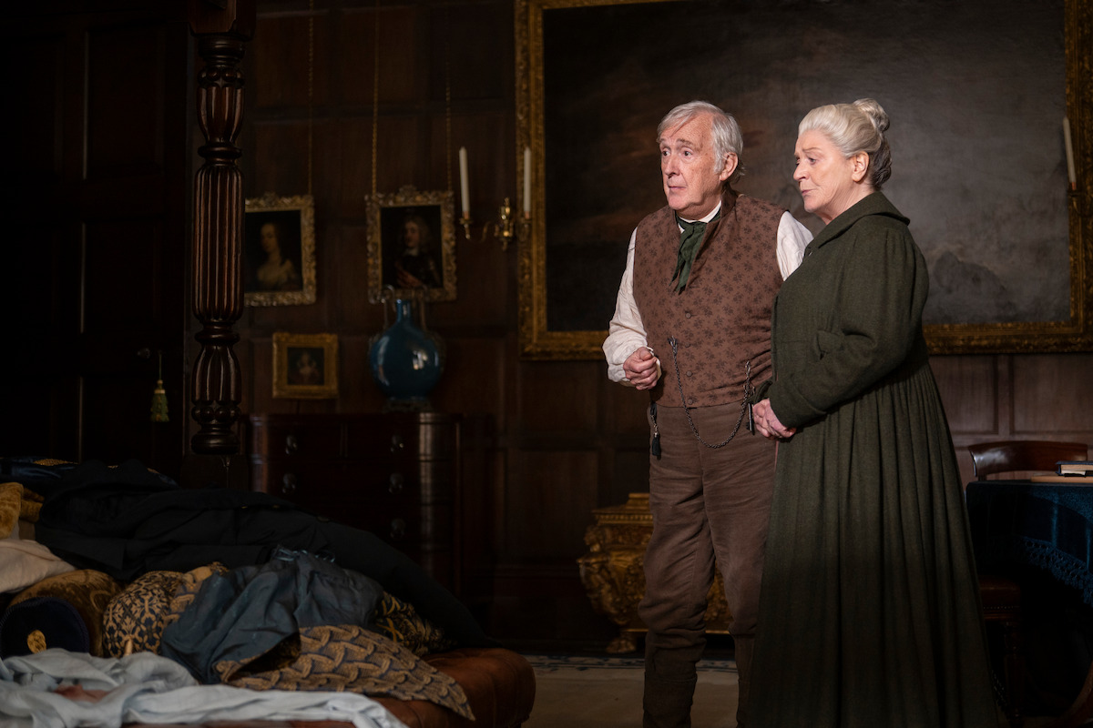 Two elderly people in period clothing stand together in a lavish, wood-paneled room with classical paintings, ornate furniture, and a bed covered with blankets and clothes in the foreground.