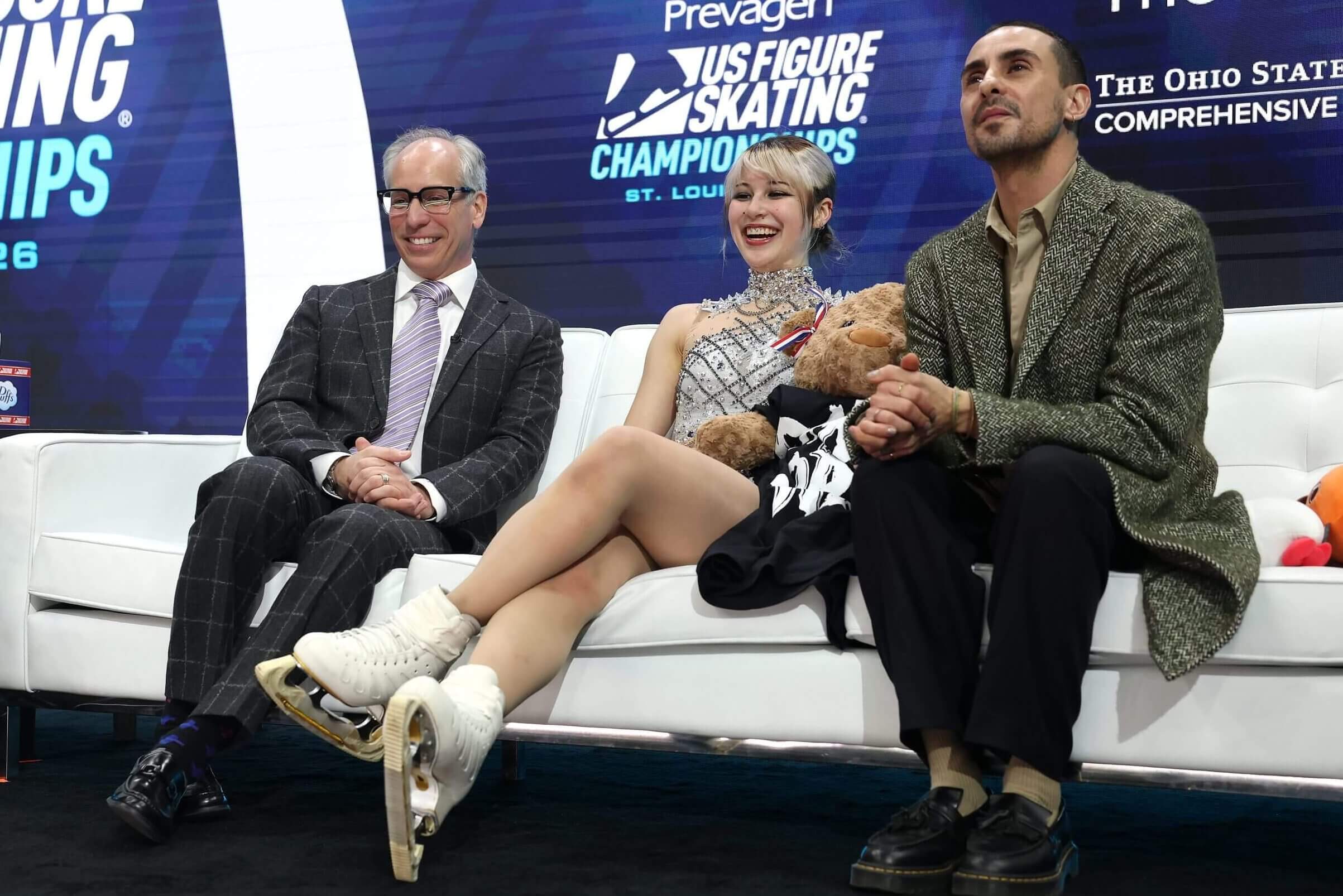 Three people seated on a white couch, with a figure skater in the middle wearing her skates.