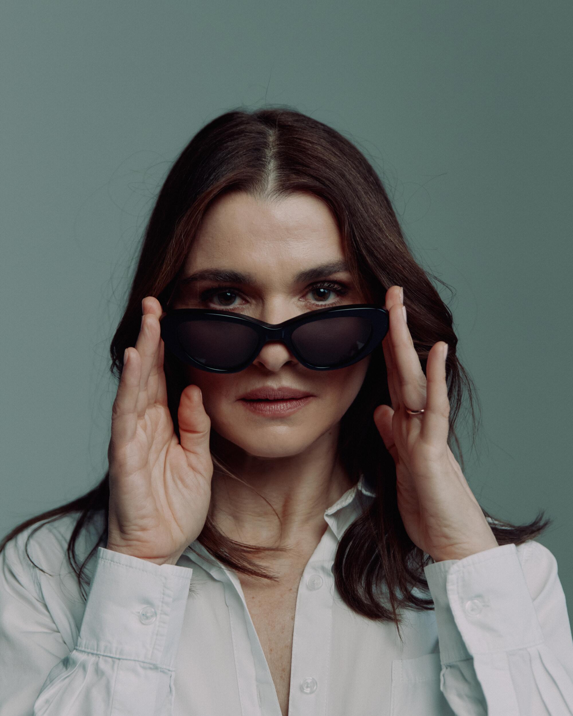 A woman with brown hair looking over a pair of sunglasses she is holding onto her face with both hands.