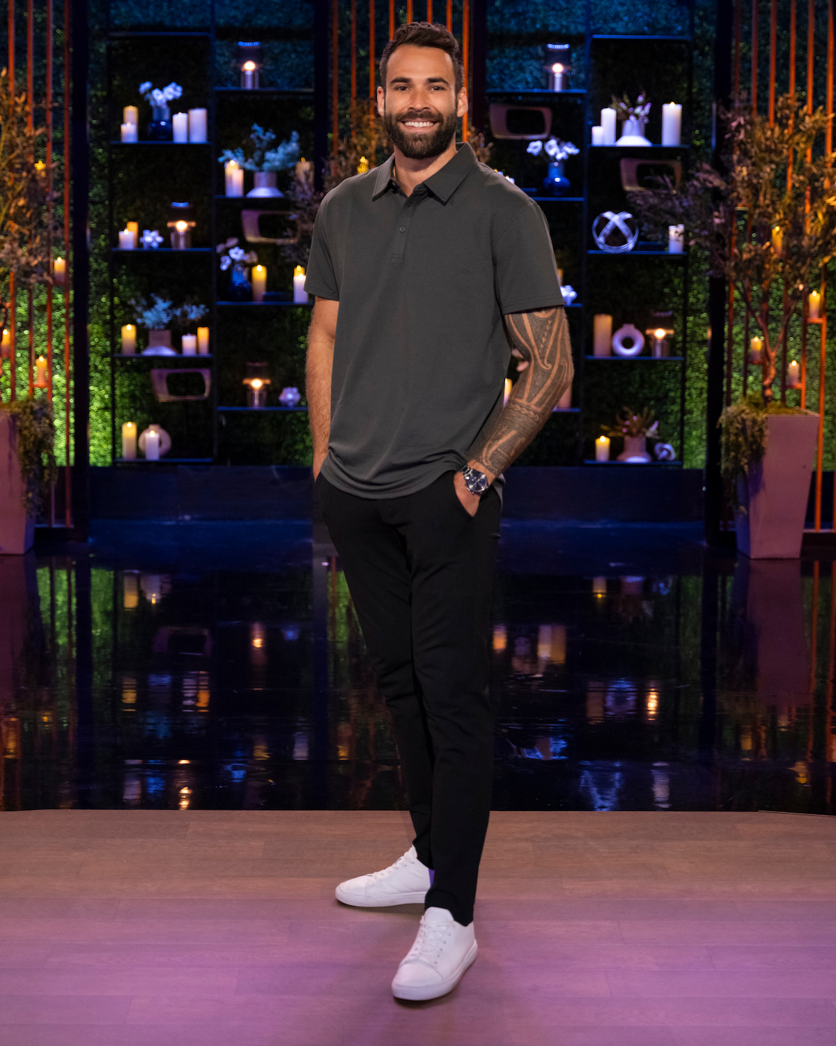 A man with a beard and tattooed arm poses indoors on a studio set decorated with candles, shelves, plants, and soft lighting, wearing a gray polo, black pants, and white sneakers, smiling at the camera.