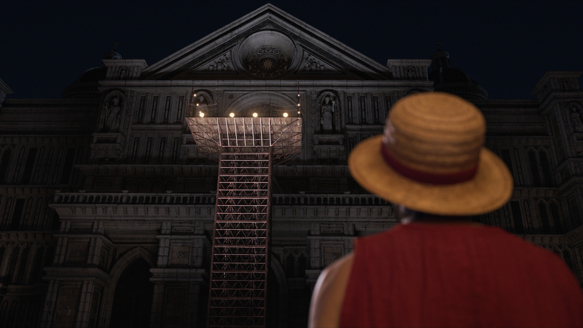 Person in a straw hat stands at night facing a grand, illuminated stone building with a scaffold and platform leading to a lit entrance above.