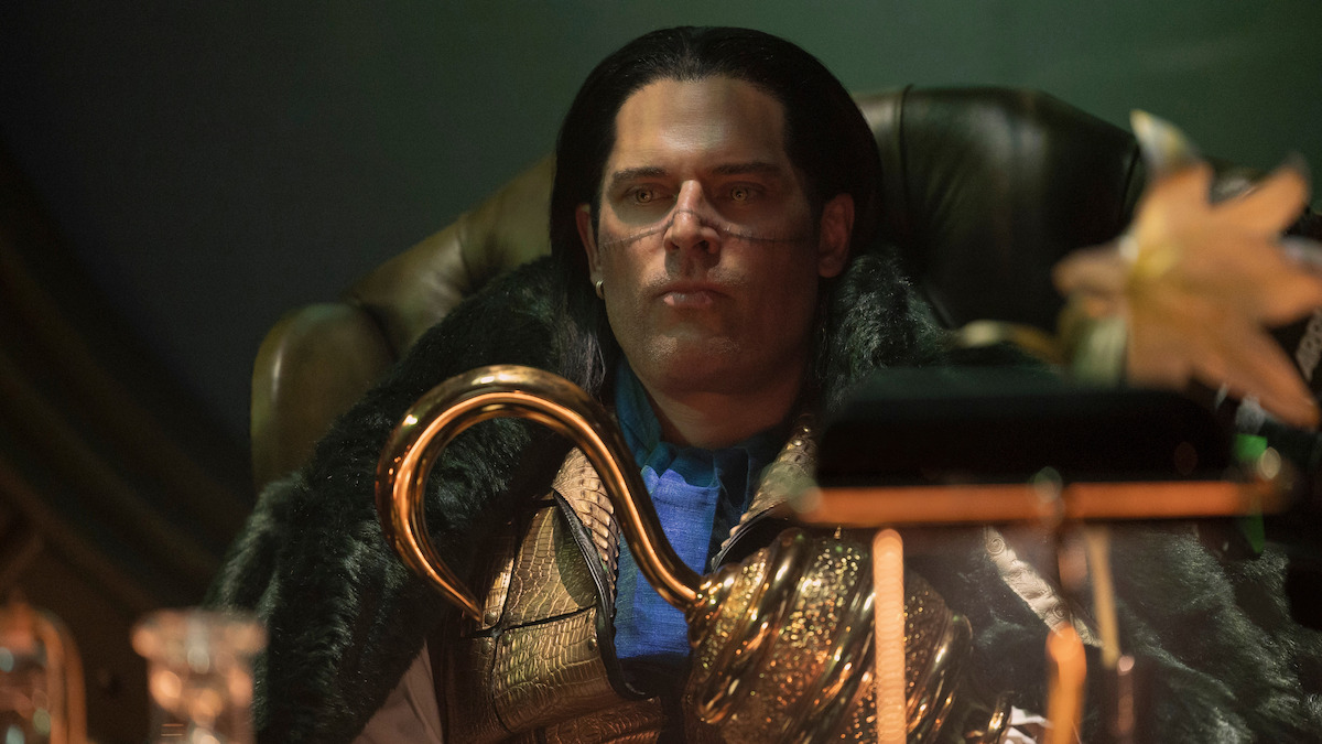 Joe Manganiello as Mr. Zero, with long dark hair sits in a dimly lit room on a leather chair, wearing glasses and a fur-collared cloak, holding a golden staff. The background is mysterious with soft, dramatic lighting and ornate decor.