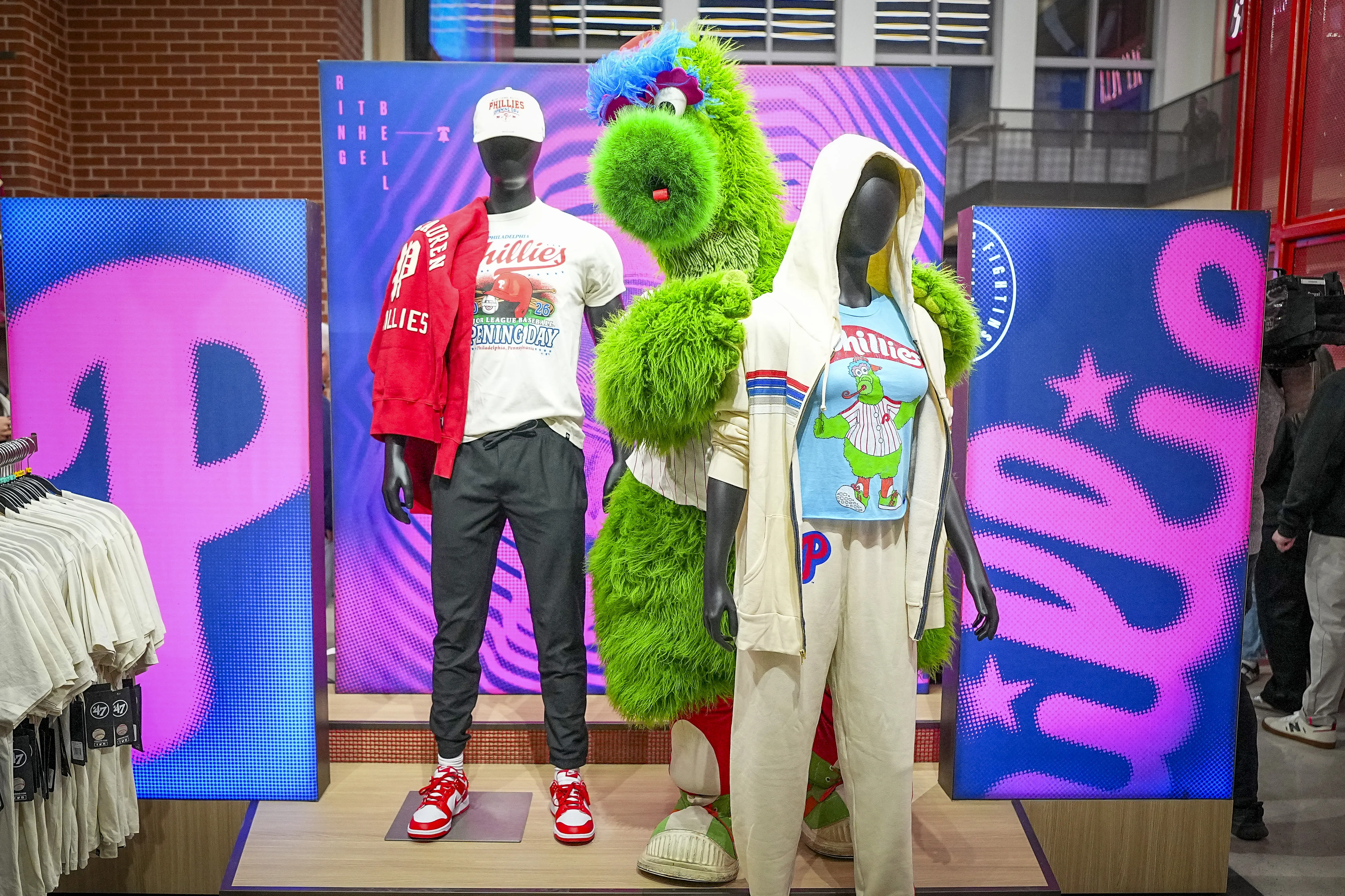 Philly-themed fashion display with two black mannequins in Phillies outfits and a bright green furry mascot behind them; pink and blue backdrops.