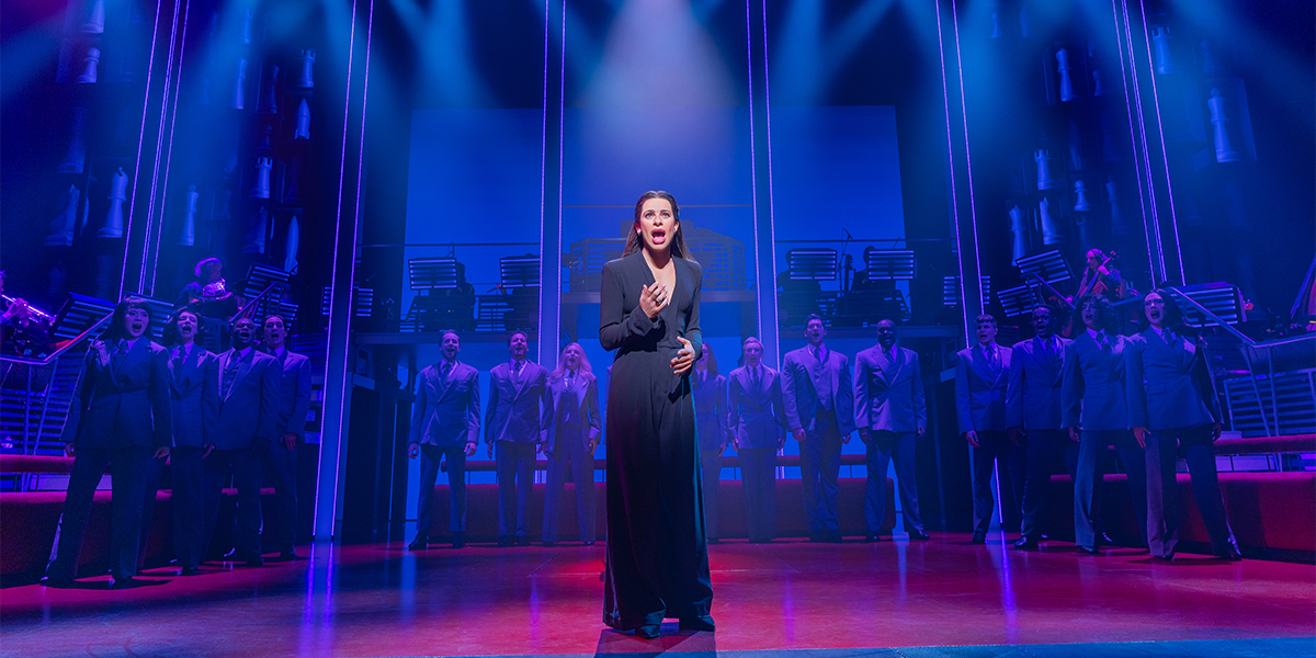 A woman stands and sings at center stage in dramatic blue and purple lighting, with a chorus of people in suits standing behind her on a theater set.