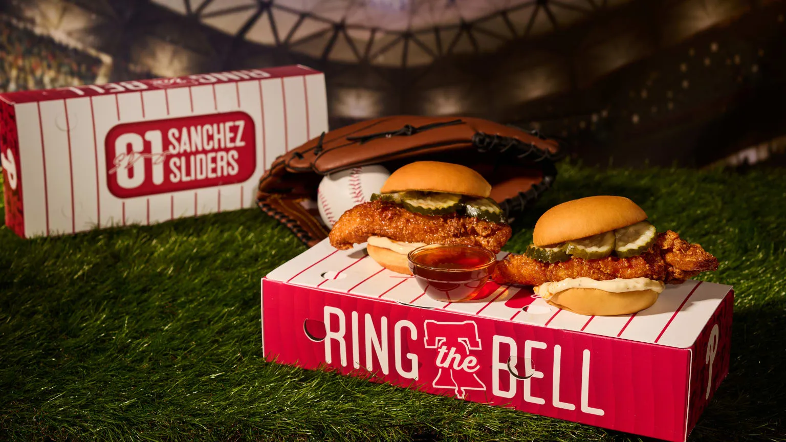 Two crispy chicken sandwiches with pickles on buns sit atop a red-and-white "Ring the Bell" box. A baseball glove and ball rest in the background, with a small sauce cup on green turf.